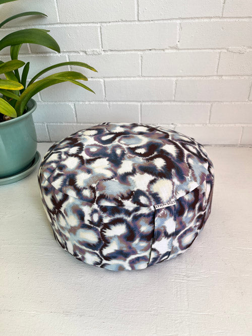 Round meditation cushion with an abstract pattern on a white surface next to a plant.