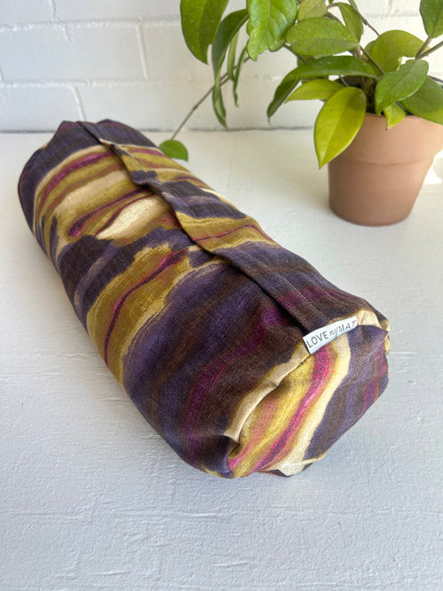 Small round yoga bolster with purple and gold pattern on a white surface with a plant in the background