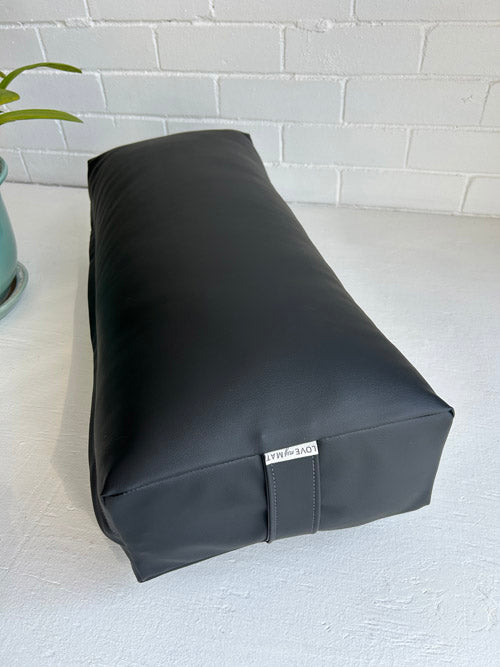 Black traditional Yoga bolster with a brand label on a white surface and light gray tiled wall background