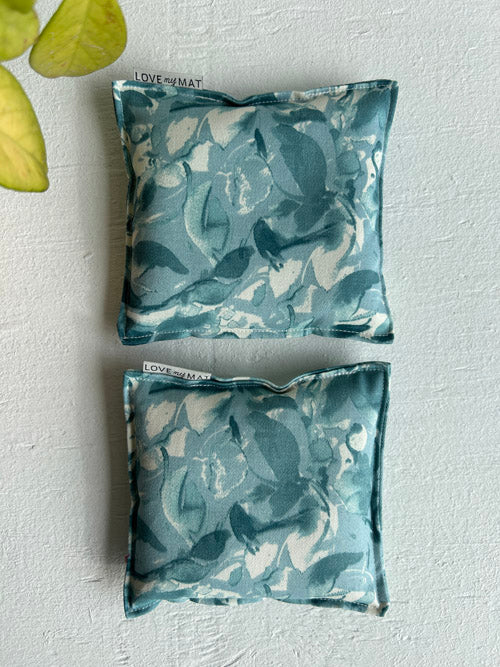 Two blue floral knee pillows on a light gray surface with a plant leaf in the corner.