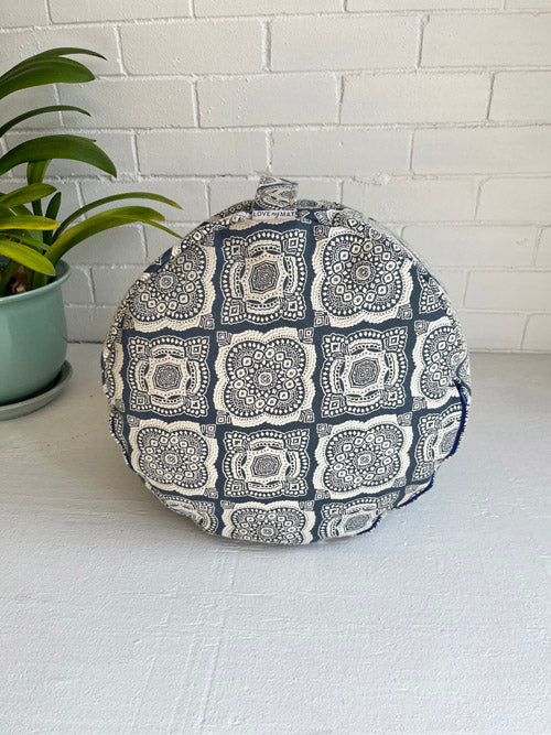 Meditation floor cushion in a blue and white geometric pattern on its side with a carry handle on top on a white surface and a white painted brick background.