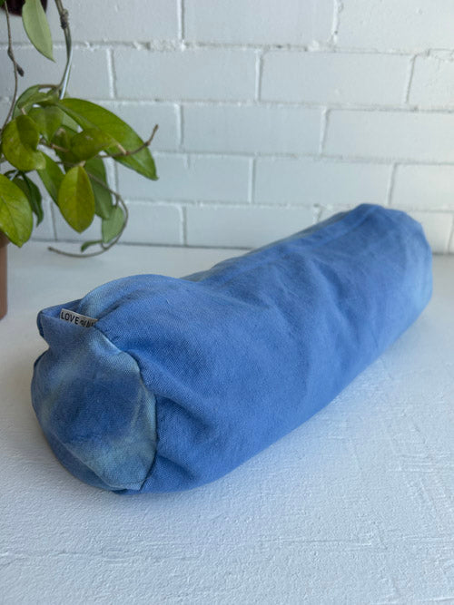 Blue cylindrical pillow on a white surface with a plant in the background