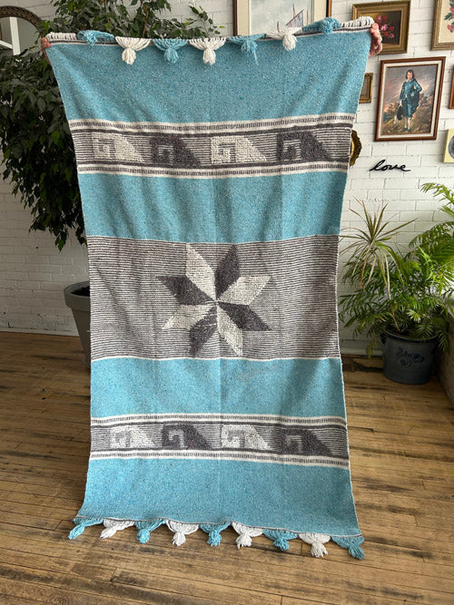 Turquoise and gray patterned blanket or tapestry  with tassels held up indoors.