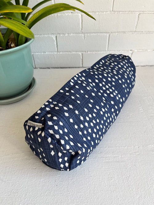 Mini round yoga bolster in blue fabric with white diamond shapes on a white surface and white painted brick background.