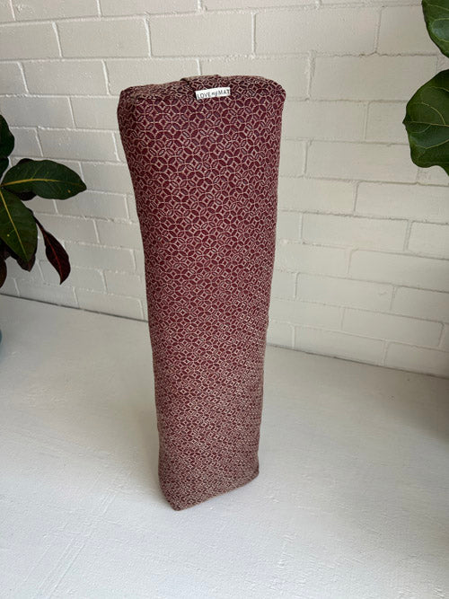 Maroon yoga bolster with pattern on a white surface against a white brick wall.