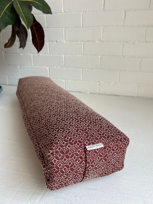 Burgundy patterned yoga bolster with a brand label on a white surface against a white brick wall.