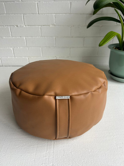 Brown leather meditation cushion with a visible brand label against a white brick wall.