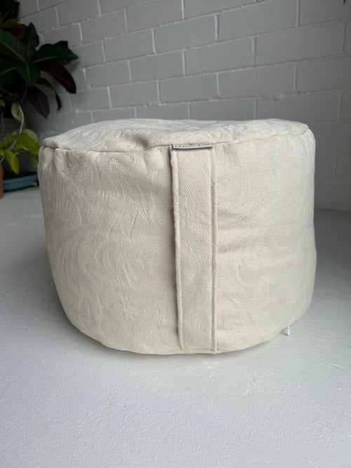 Beige fabric meditation cushion on a light gray surface with a white brick wall background