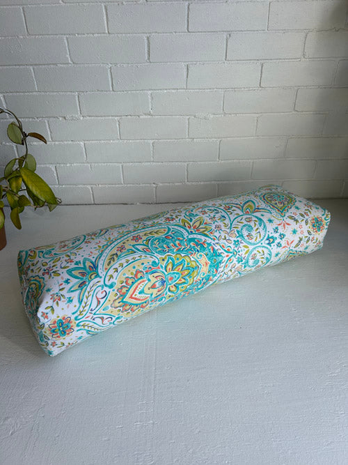 Decorative long yoga bolster with a colorful pattern on a light gray surface against a white brick wall.