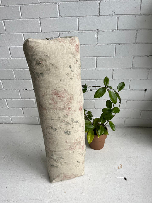A beige yoga bolster with a floral pattern, placed next to a small potted plant on a white surface against a white brick wall.