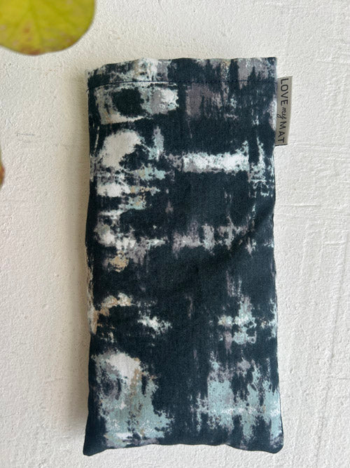 Abstract green and grey patterned eye pillow with a visible brand tag on a textured white background