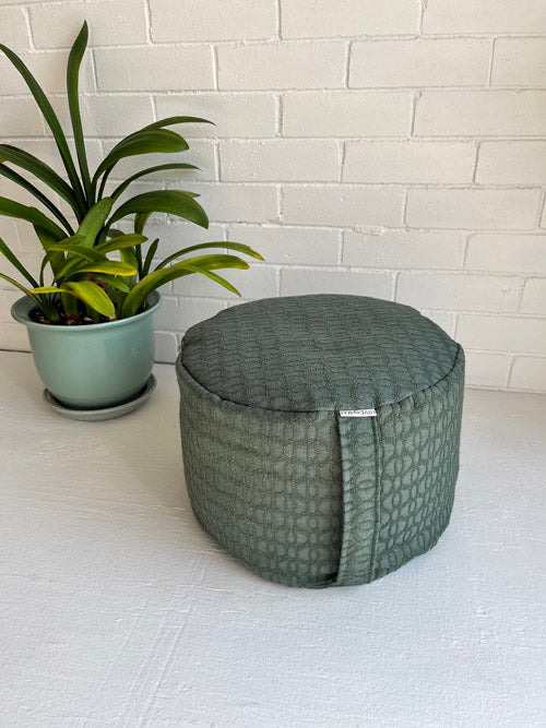 Green textured tall meditation floor cushion next to a potted plant against a white brick wall.