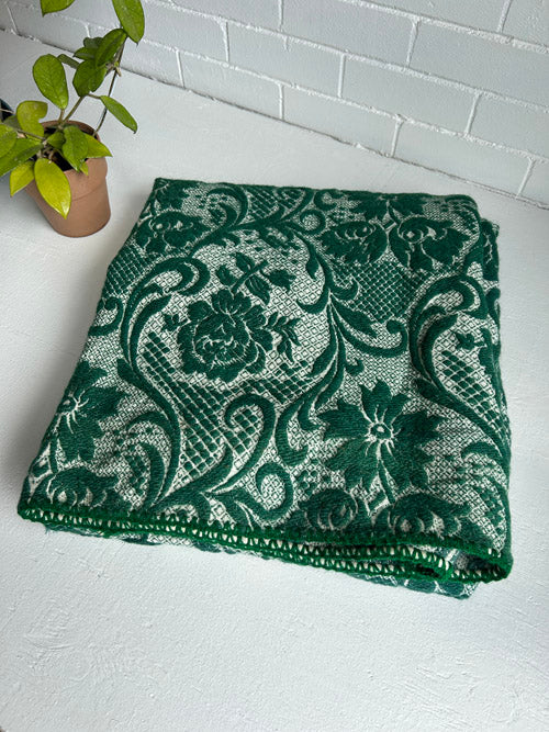 Green floral patterned wool tapestry on a white surface with a plant in the background
