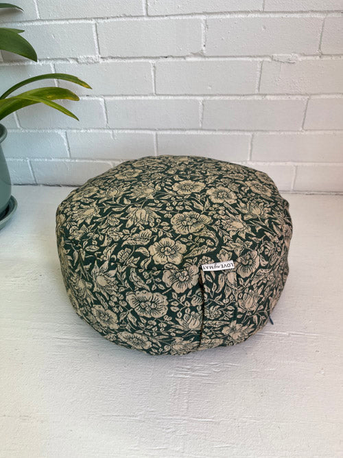 Floral-patterned meditation cushion on a white surface with a white tiled wall in the background and plant near it.