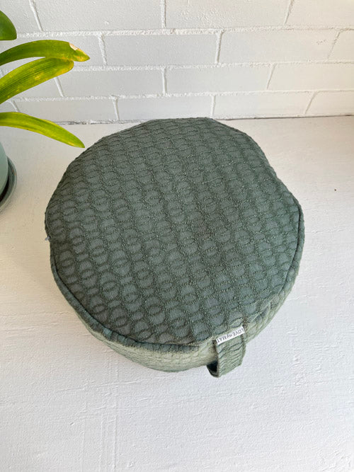 Green textured meditation floor cushion on a white surface with a plant in the corner