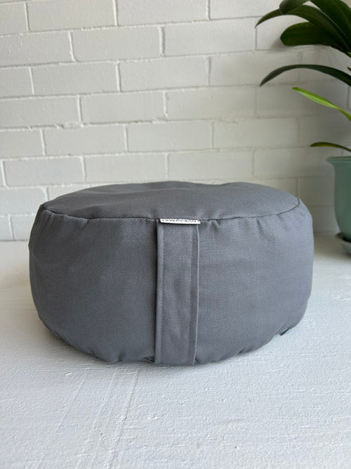 Gray meditation cushion on a light gray floor with a white brick wall in the background.