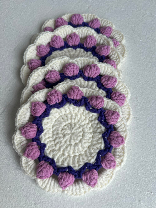 Set of white crocheted coasters with purple pom-pom trim on a gray background