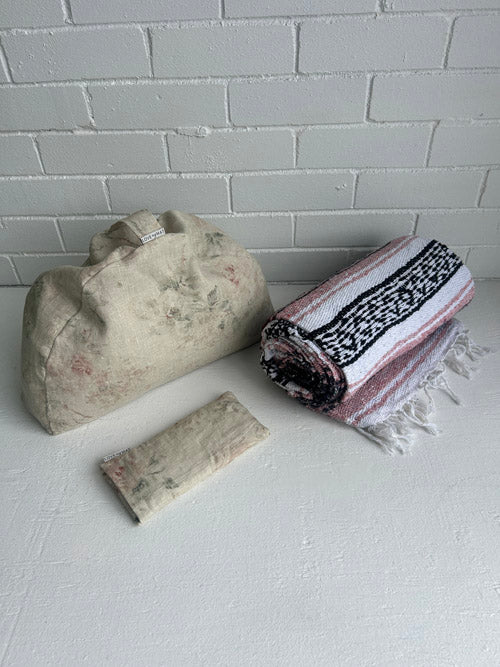 Fabric sustainable meditation cushion, folded pink and black blanket, and fabric eye pillow on a white surface with a white painted brick background.