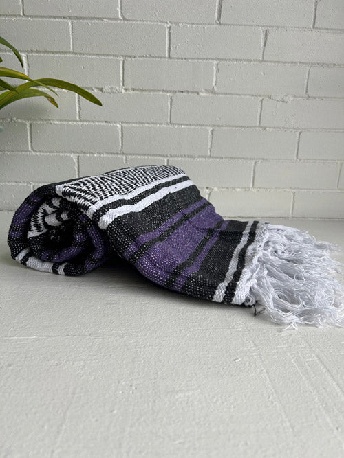Rolled-up purple and black striped Mexican blanket with white tassels on a light gray surface.