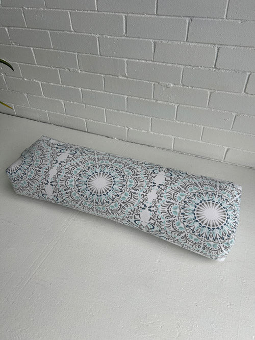 Long yoga Bolster with mandala design on a light gray surface against a white brick wall.