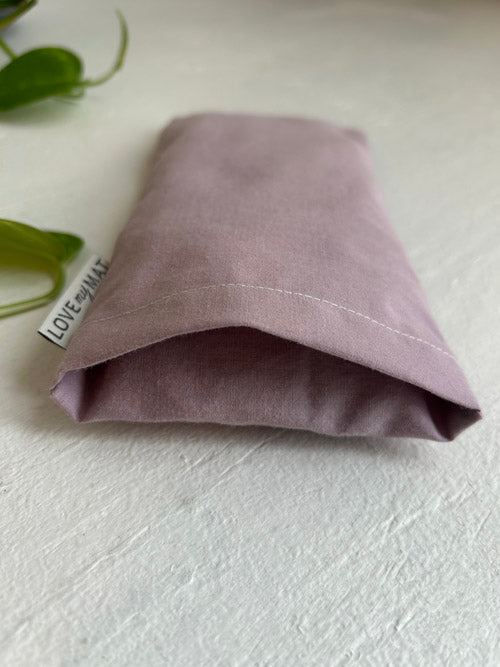 Folded purple fabric eye pillow on a light gray surface with green leaves in the corner.