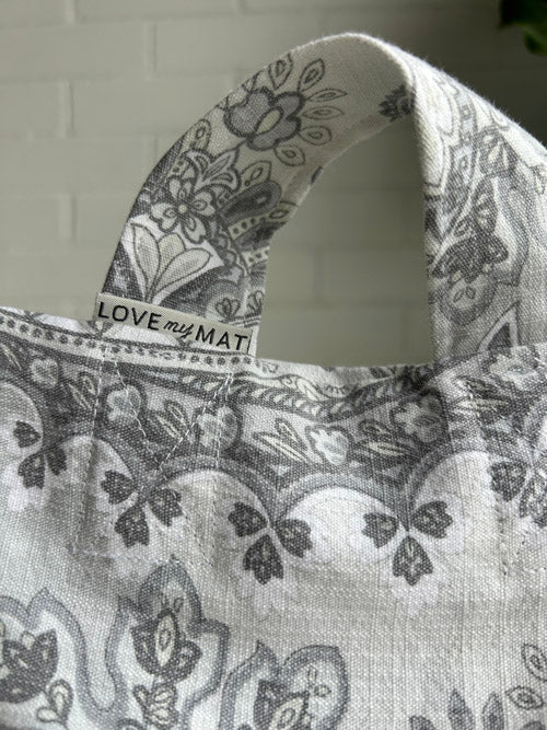 Gray patterned tote bag with 'LOVE my MAT' label on handle against a white brick wall.