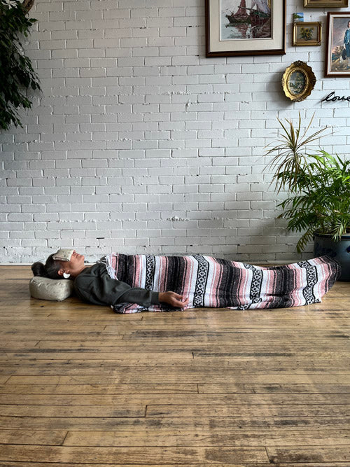Person lying on a meditation cushion, an eye pillow on eyes and a striped blanket over them with a white brick wall and plants in the background.
