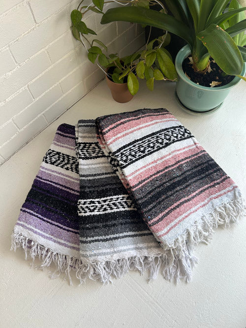 Woven blanket with fringe on a white surface with plants in the background