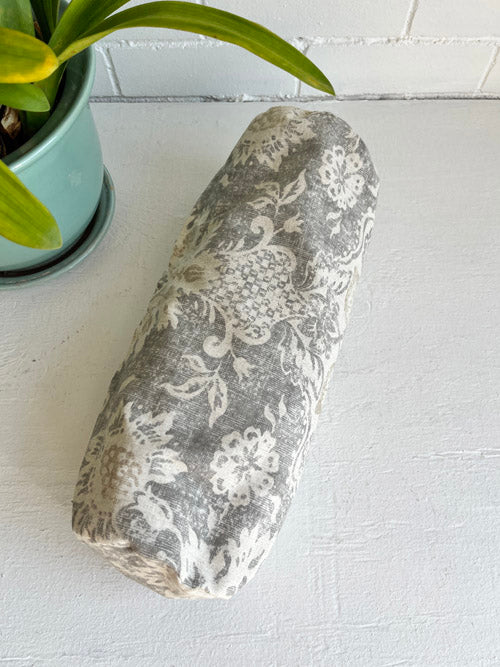 Neutral floral patterned mini round yoga bolster on a white surface next to a potted plant.