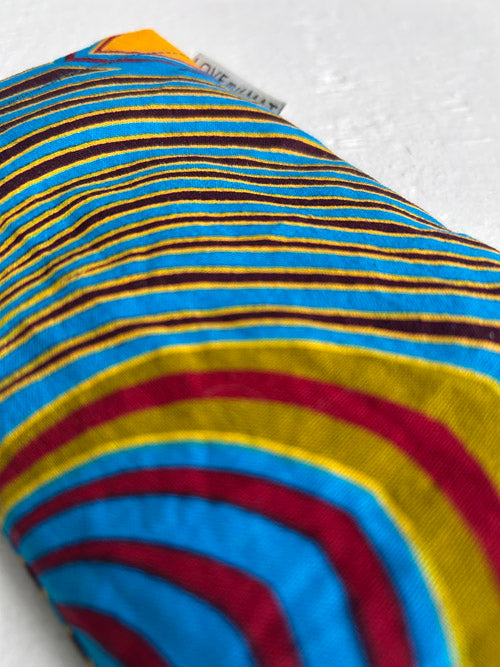 Close-up of a colorful striped fabric eye pillow with blue, yellow, and red patterns on a white background.