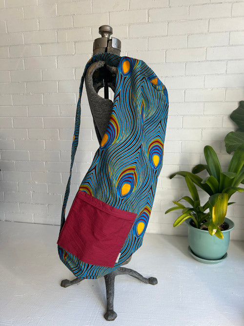 Colorful fabric yoga mat bag with peacock feather pattern on a mannequin against a white brick wall.