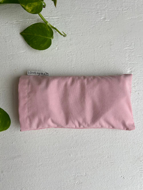 Pink fabric eye pillow on a light gray background with green leaves.