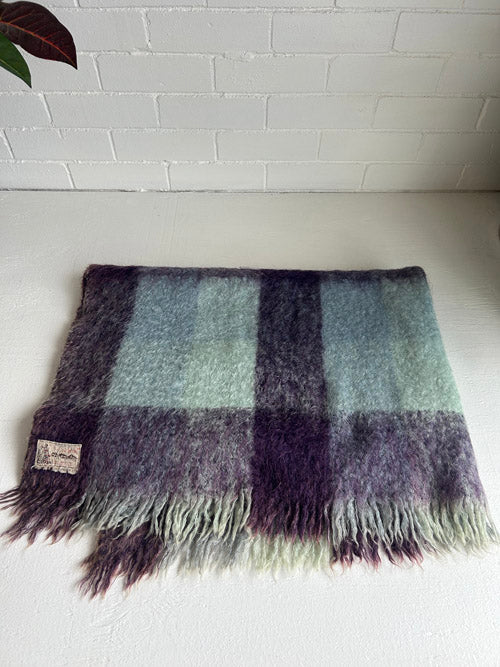 Plaid blanket with fringes on a white surface against a white brick wall.