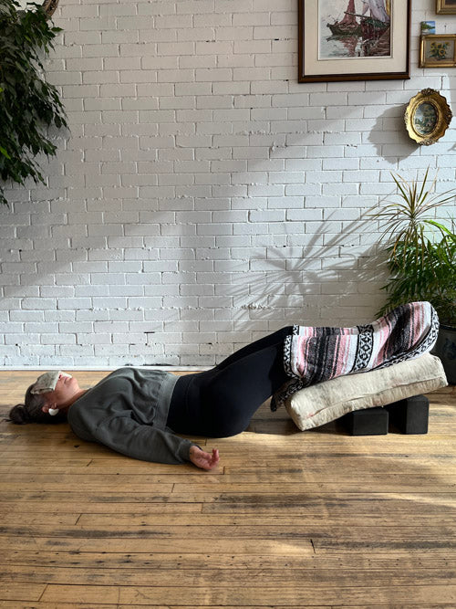 Person lying down with an eye pillow on eyes, legs covered in a blanket and propped up using a bolster and yoga blocks in a room with a white brick wall and plants.