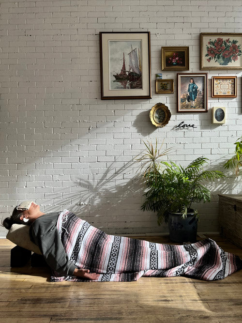 Person lying back on a yoga bolster propped up with yoga blocks, an eye pillow on eyes and a striped blanket over them in a room with white brick walls and framed pictures.
