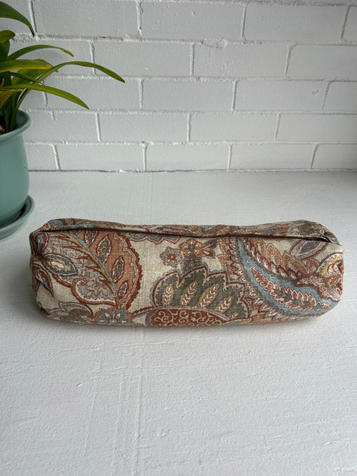 Fabric small round bolster with paisley pattern on a white surface with a plant in the background