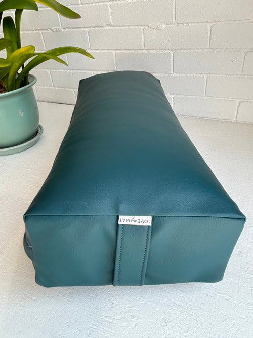 Teal vinyl yoga bolster with a brand label on a light surface next to a plant.