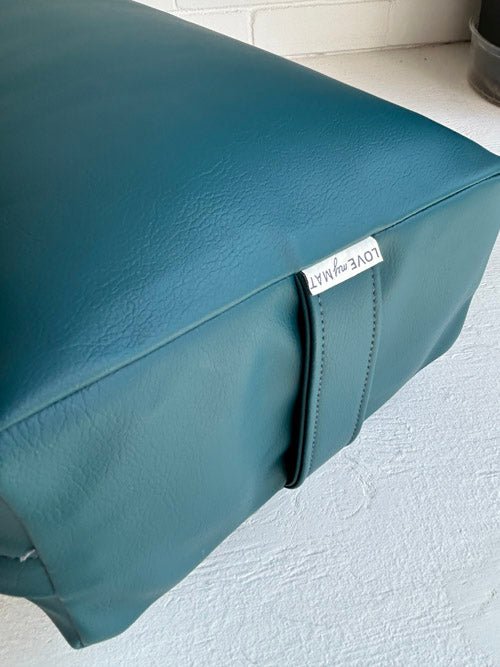 Teal leather ottoman with a brand label on a light gray surface