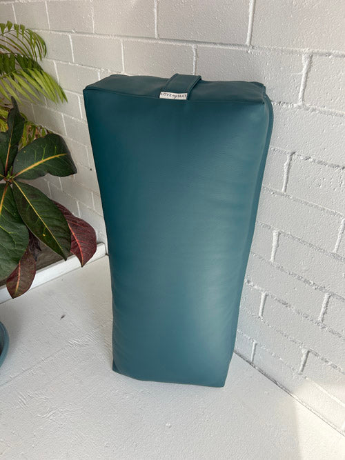 Traditional Yoga Bolster in teal vinyl with a brand label on a white surface next to a plant