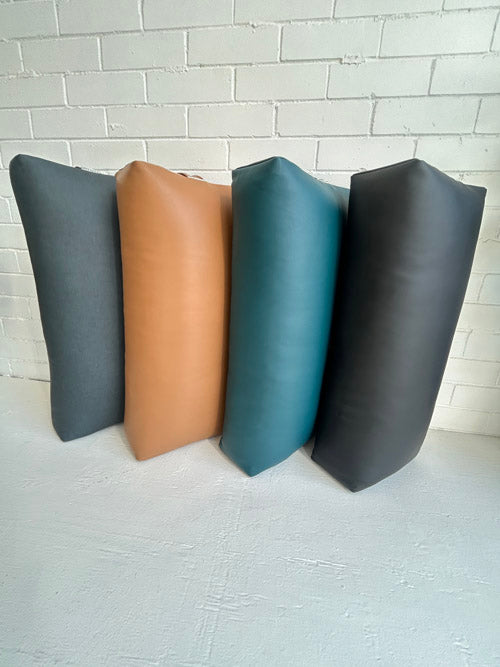 Four leather yoga bolster in gray, brown, blue, and black on a light gray surface with a white brick wall background.