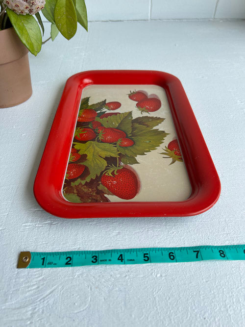 Red tin tray with strawberry design on a white surface, measured by a tape measure.