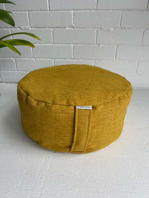 Mustard yellow meditation cushion with a brand tag 'LOVE my MAT' on a white surface and white brick wall background