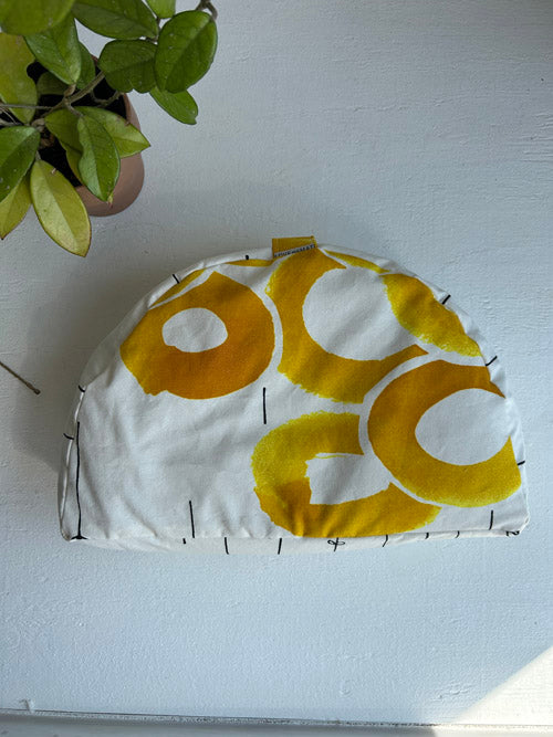 White with yellow floral pattern sit pillow on a white surface with a plant in the corner.
